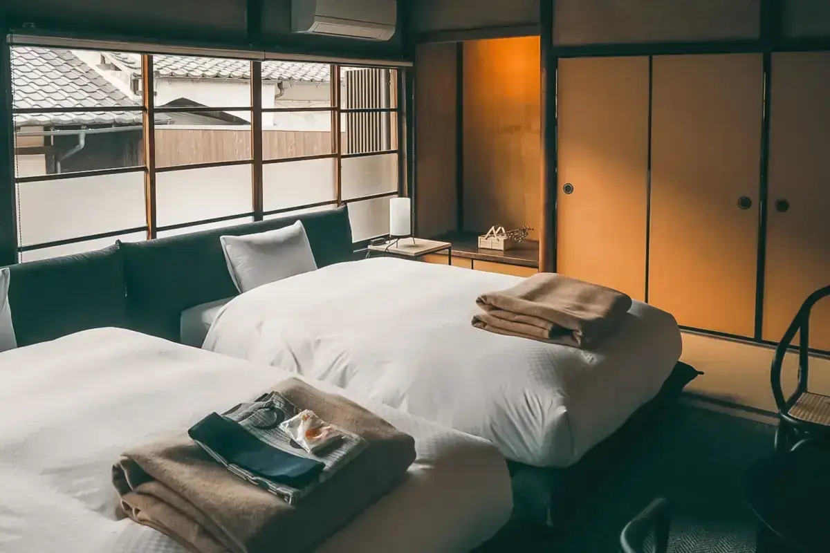Traditional Japanese hotel room with twin beds featuring white linens and folded towels, sliding shoji doors, and windows overlooking tiled rooftops.