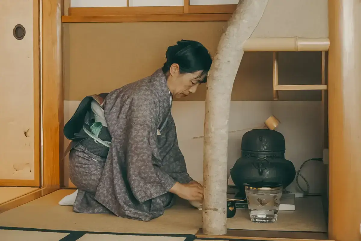 Woman in traditional gray kimono kneeling in formal seiza position during Japanese tea ceremony, preparing tea in a traditional tatami room with a cast iron kettle and tokonoma alcove.
