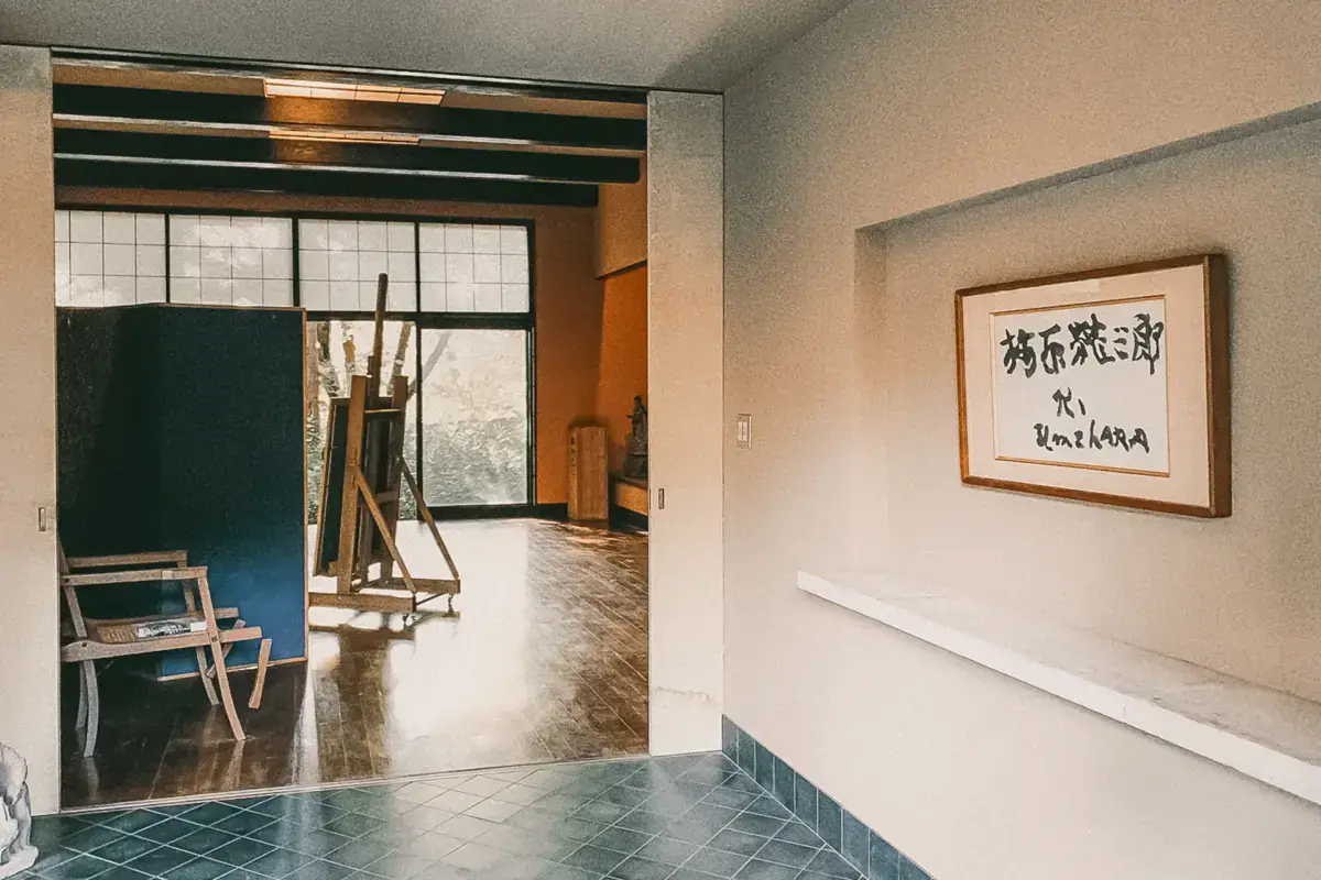 Traditional Japanese interior entrance with green-tiled floor, framed calligraphy on the wall, and a view into an art studio with easel and furniture featuring grid-patterned windows.