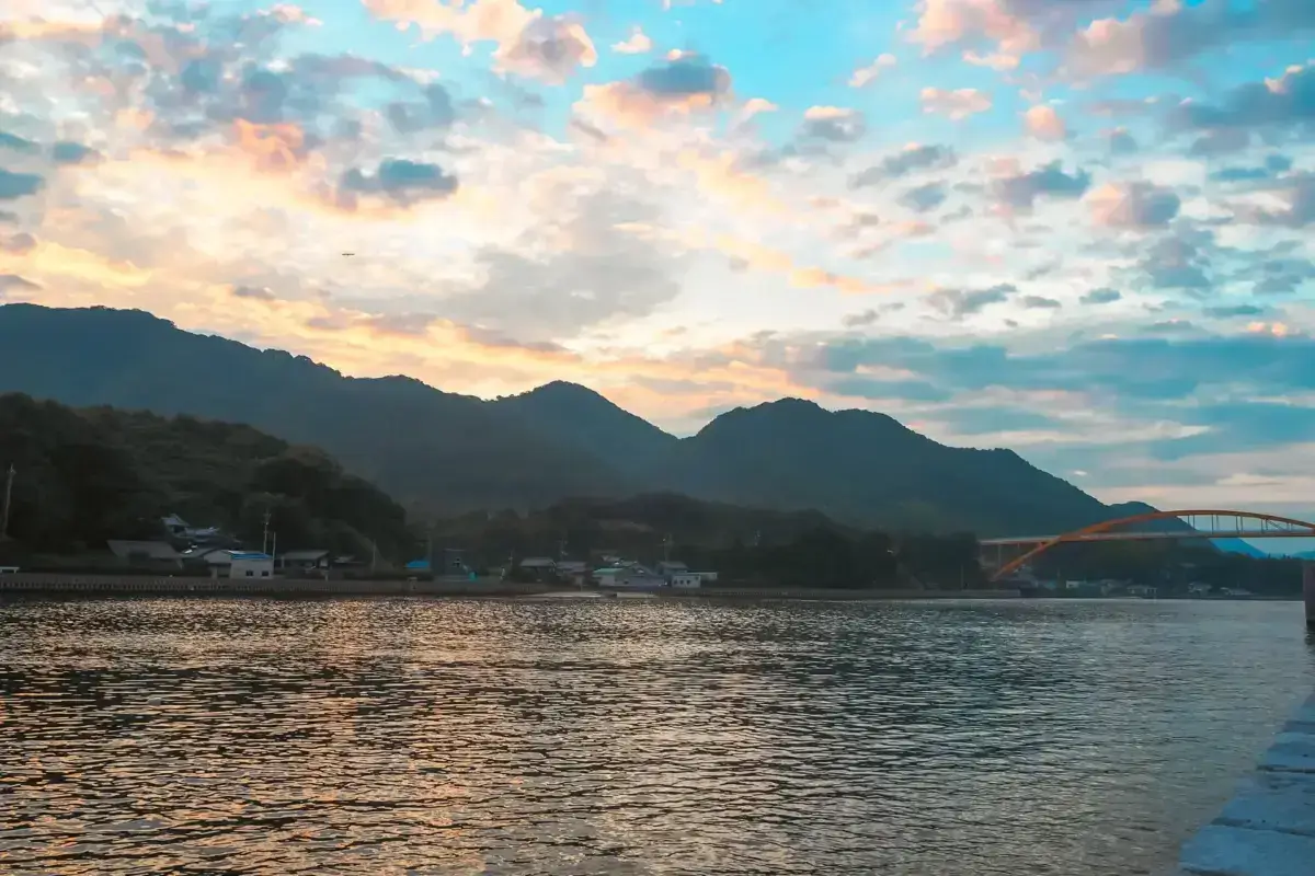Sunset view over a calm river with silhouetted mountains and a small coastal town with an arched bridge, featuring dramatic clouds in shades of orange, pink, and blue.