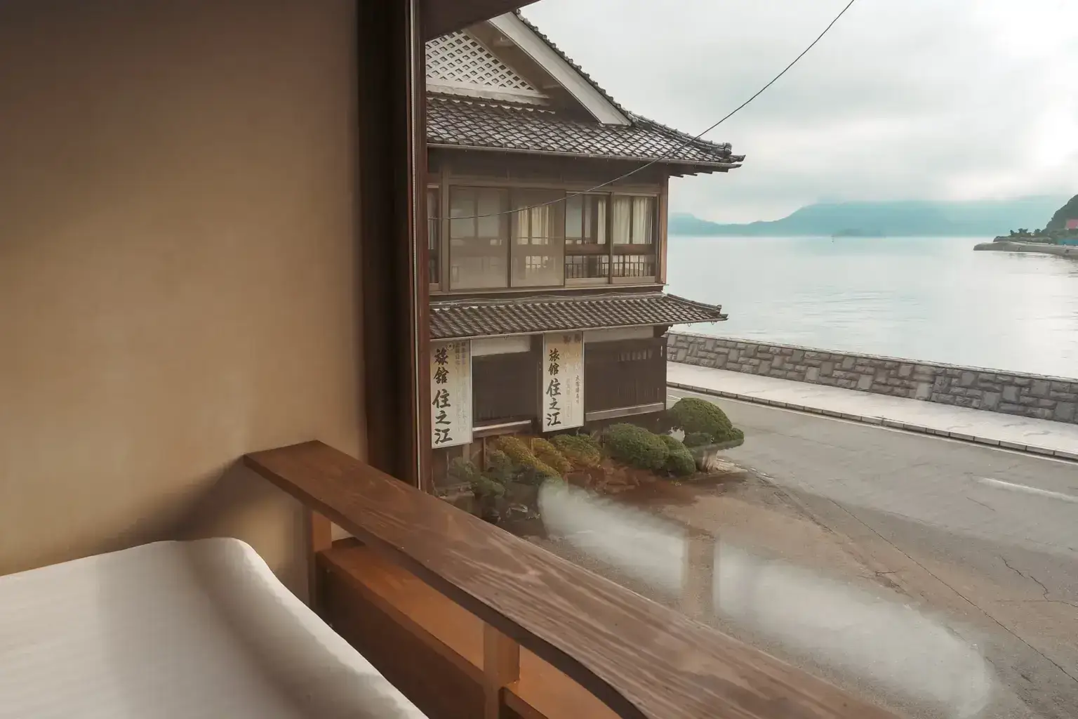 Traditional Japanese inn window view overlooking a tranquil waterfront with tiled-roof buildings, manicured gardens, and misty mountains in the background.
