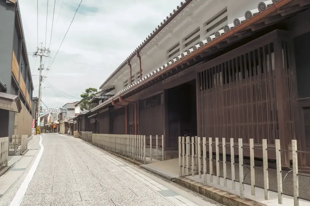 Traditional Japanese street with wooden machiya buildings featuring dark lattice facades and ornate tiled roofs, lined by a low wooden fence along a quiet stone-paved walkway.