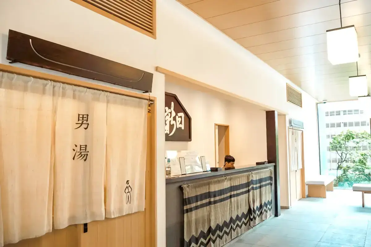 Japanese bathhouse reception area with noren curtain entrance on the left displaying kanji characters for "men's bath" and a front desk with traditional textile decoration and staff member in the center.
