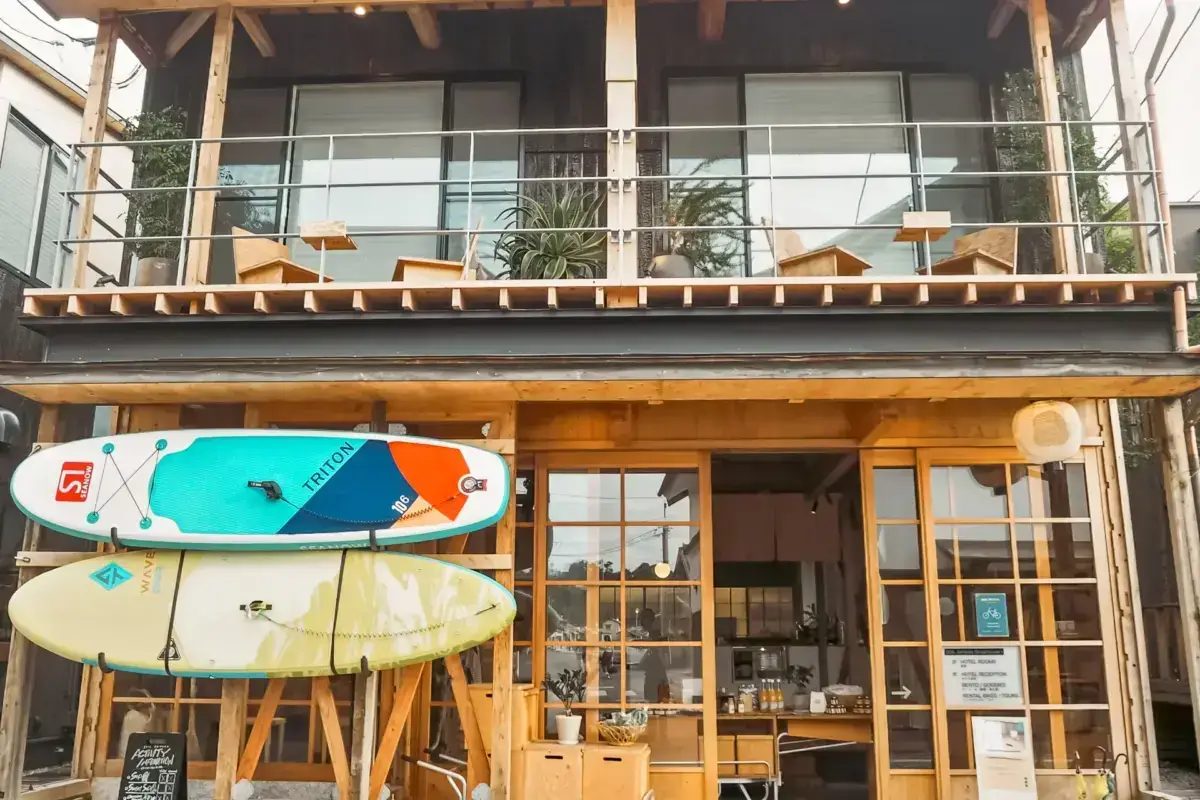 Two-story modern beach house under construction with wooden frame balcony and three surfboards displayed on the ground floor storefront entrance.