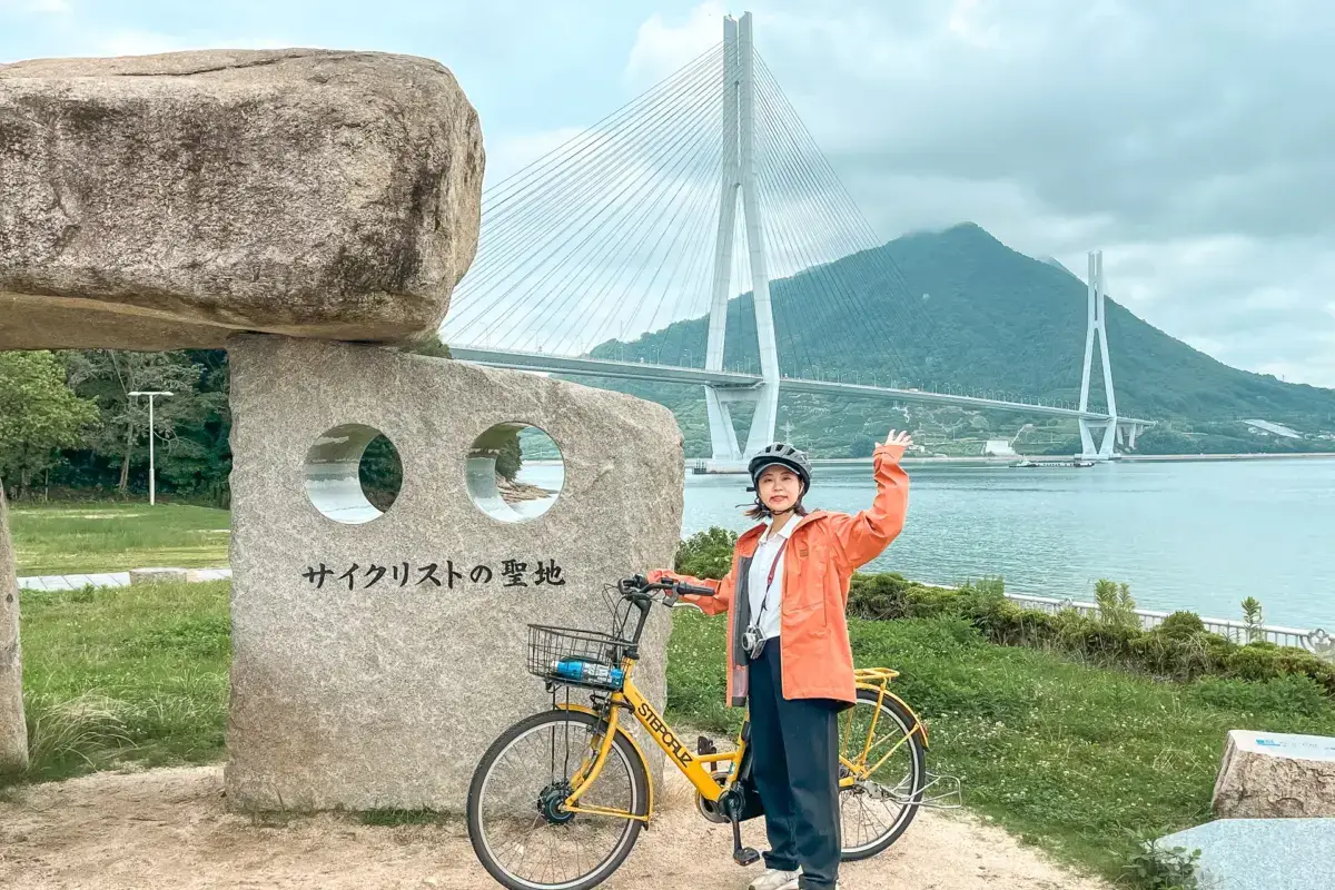 Woman with bicycle standing beside stone monument with Japanese text at waterfront park, featuring modern cable-stayed bridge and mountain backdrop on Shimanami Kaido cycling route.
