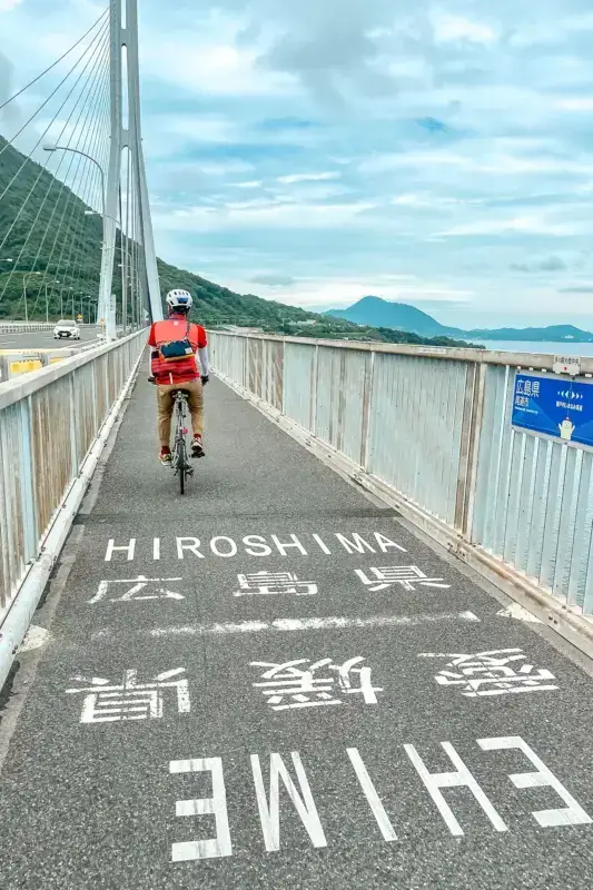 Cyclist in red jacket riding along Shimanami Kaido bridge bike path with cable-stayed bridge structure on left and scenic ocean views of Japanese islands in the background under partly cloudy skies