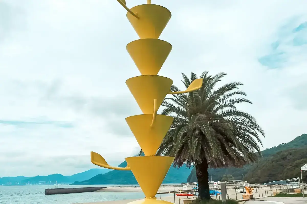 Modern yellow sculptural tower with stacked conical shapes beside a palm tree at a waterfront promenade with mountains in the background.