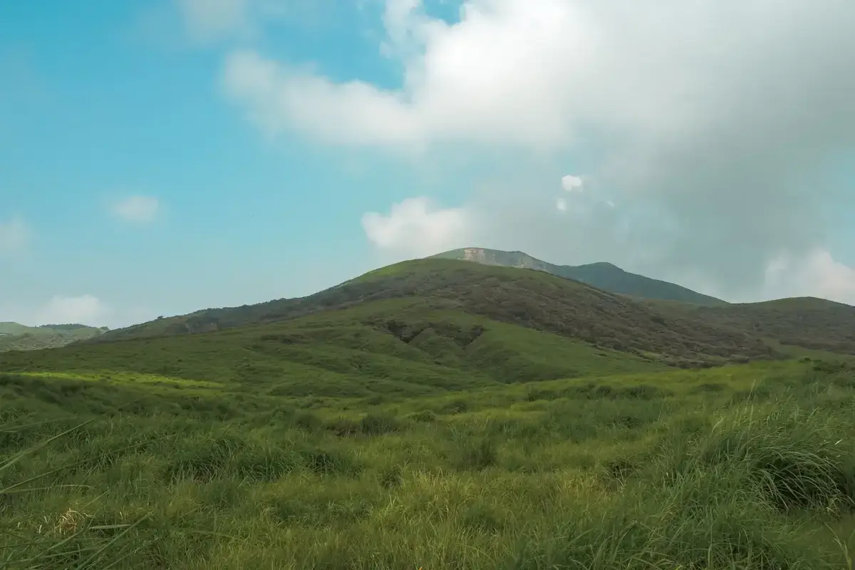 Rolling green hills and volcanic mountains under a cloudy sky in Kyushu, with lush grasslands in the foreground