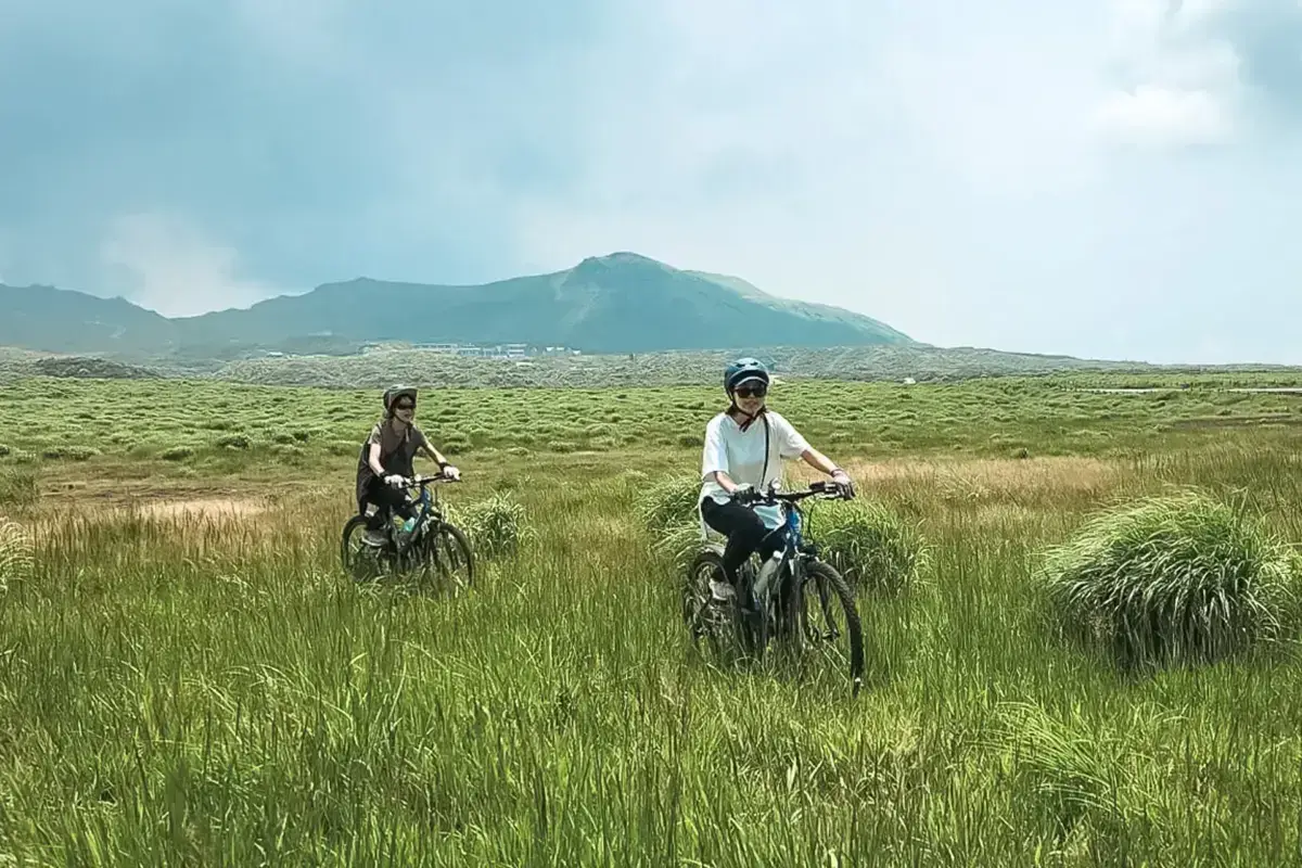 Two cyclists riding through expansive green grasslands with Mount Aso volcano rising in the background under a partly cloudy sky in Kyushu, Japan.