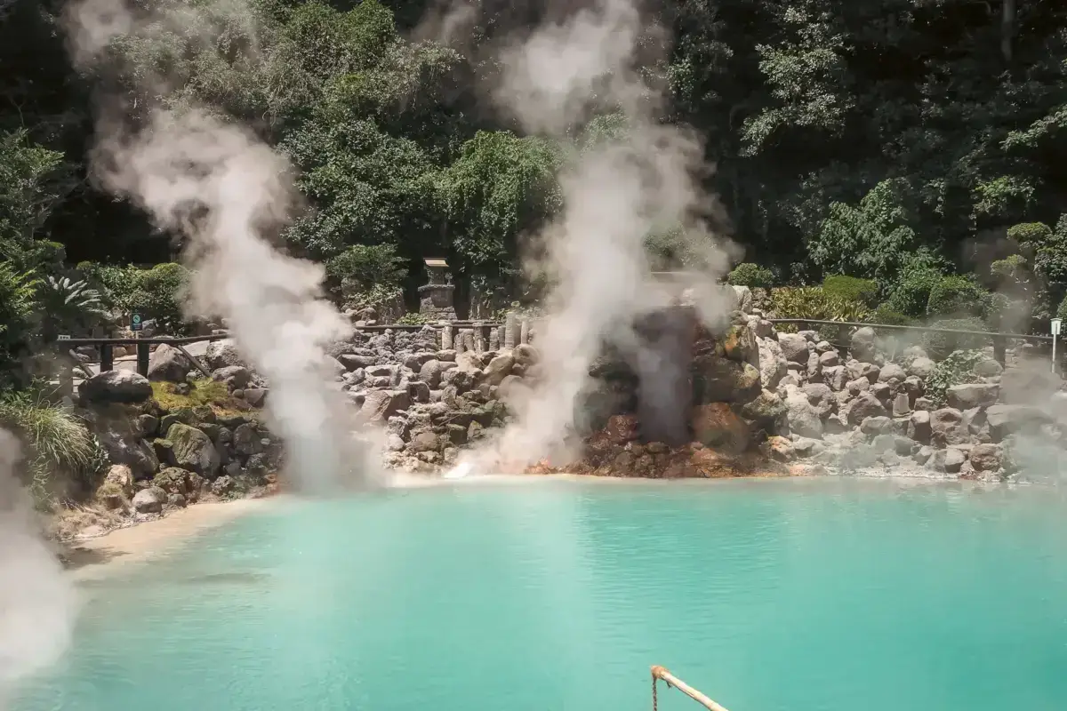 Turquoise hot spring pool with dramatic volcanic steam plumes rising from rocky geothermal vents, surrounded by lush green forest in Kyushu, Japan.