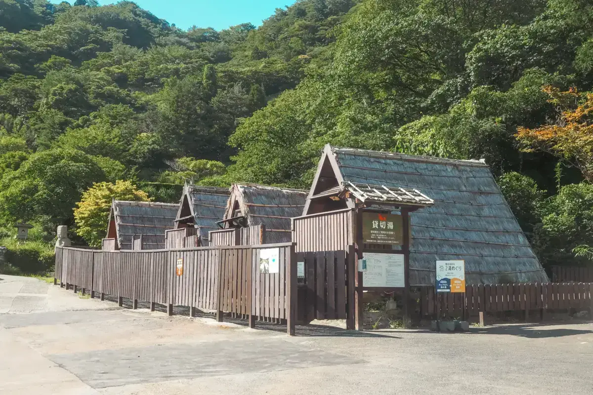 Traditional Japanese wooden structures with steep tiled roofs nestled against a lush green mountainside in Kyushu, featuring rustic timber architecture surrounded by dense forest vegetation.