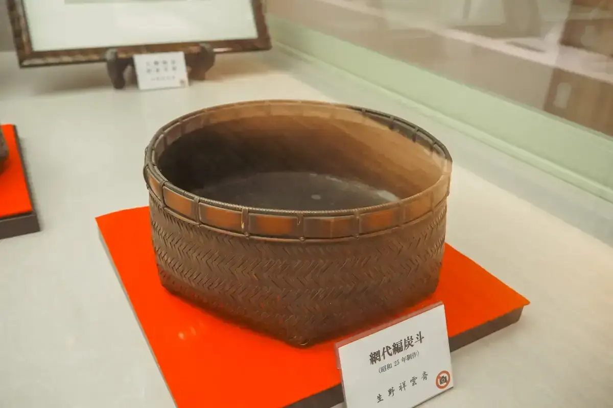 Traditional Japanese bamboo charcoal basket with woven herringbone pattern displayed on an orange platform in a museum exhibition