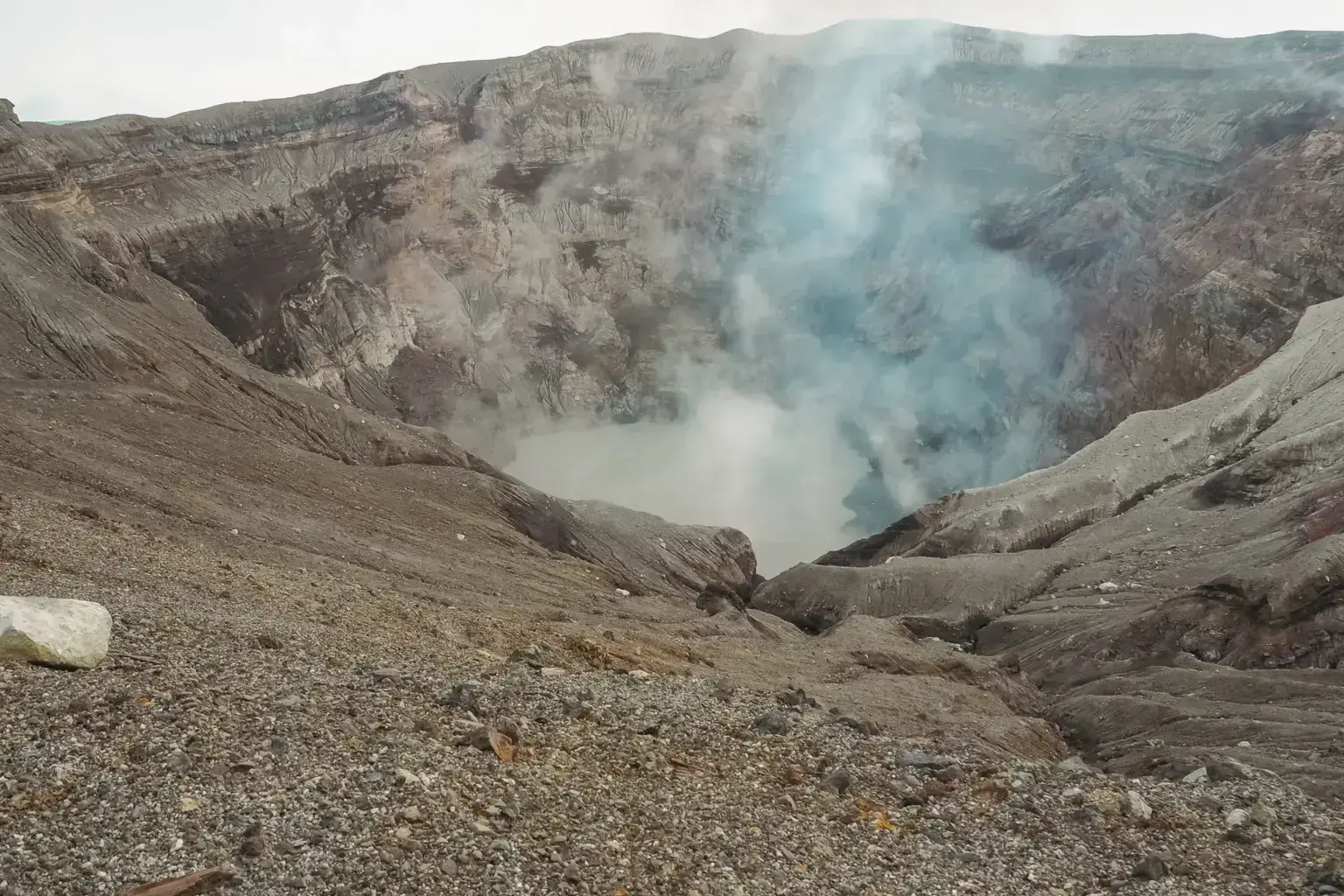 Active volcanic crater in Kyushu with steam and sulfurous gas rising from rocky slopes and a turquoise acidic lake at the bottom.