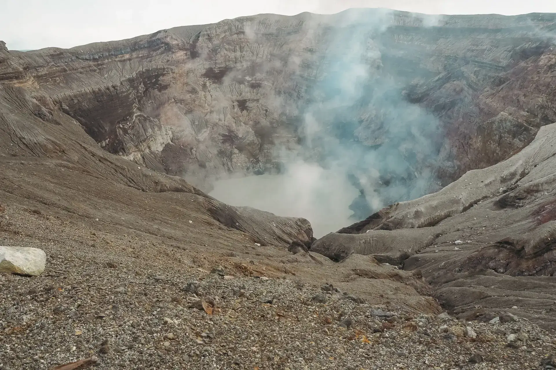 Active volcanic crater in Kyushu with steam and sulfurous gas rising from rocky slopes and a turquoise acidic lake at the bottom.