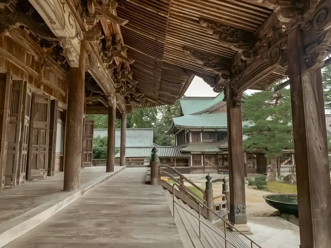 Traditional Japanese temple wooden corridor with ornate carved brackets overlooking a courtyard with buildings featuring copper-green roofs and pine trees in the Hokuriku region.