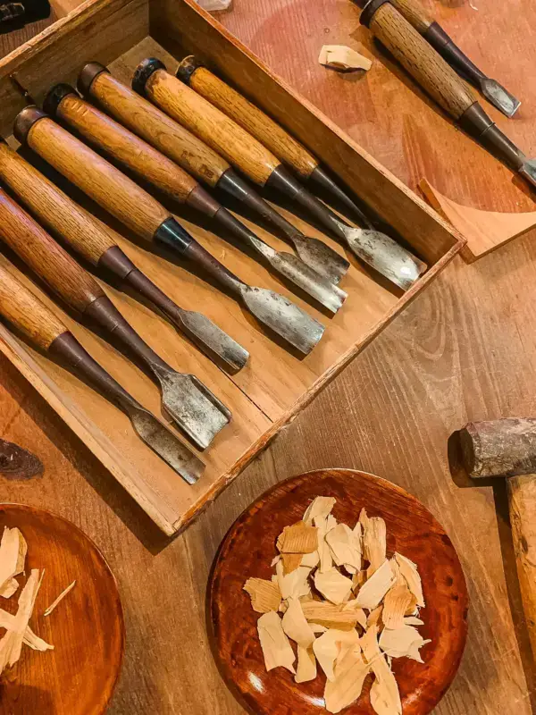 Traditional Japanese woodworking chisels with wooden handles displayed in an open case, alongside carved wood shavings in a wooden bowl and other carpentry tools on a workbench.