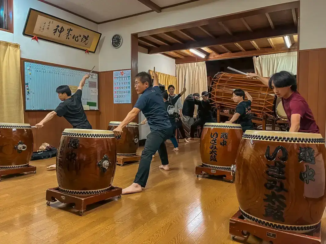 Group of people practicing taiko drumming in a traditional Japanese community hall, with drummers positioned at large wooden drums and a large odaiko drum visible in the background.