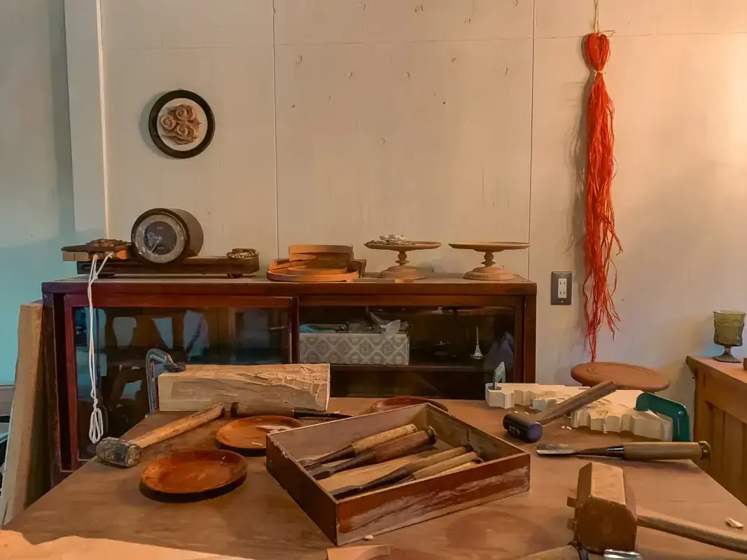 Traditional Japanese woodworking workshop with vintage wooden tools, storage cabinet displaying circular molds and wooden plates, and an orange decorative tassel hanging on the white wall.