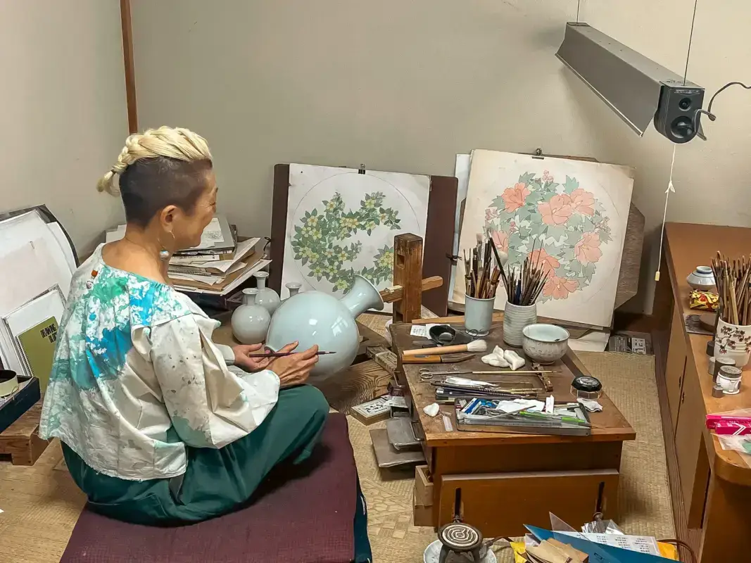 Japanese artist painting traditional floral designs on ceramic spheres in a studio workspace filled with art supplies, brushes, and completed botanical artworks displayed on easels.