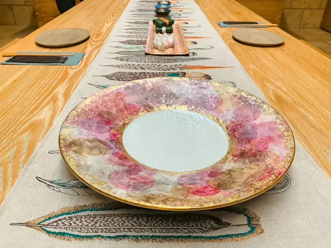 Elegant Kutani porcelain plate with colorful floral pattern and gold rim placed on decorative table runner, part of a traditional Japanese table setting with wooden table and matching place settings in the background.