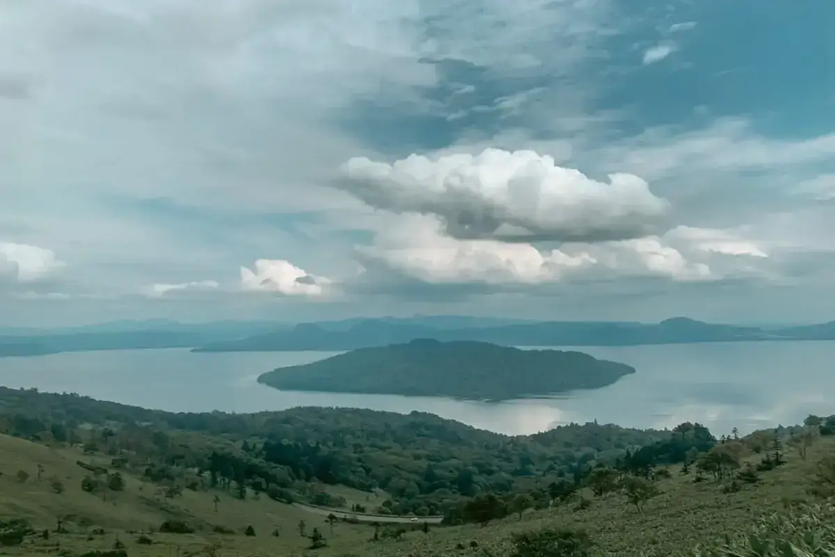 Panoramic view of Lake Kussharo in Teshikaga, Hokkaido, featuring a forested island surrounded by calm blue waters with rolling hills in the foreground and dramatic clouds overhead.