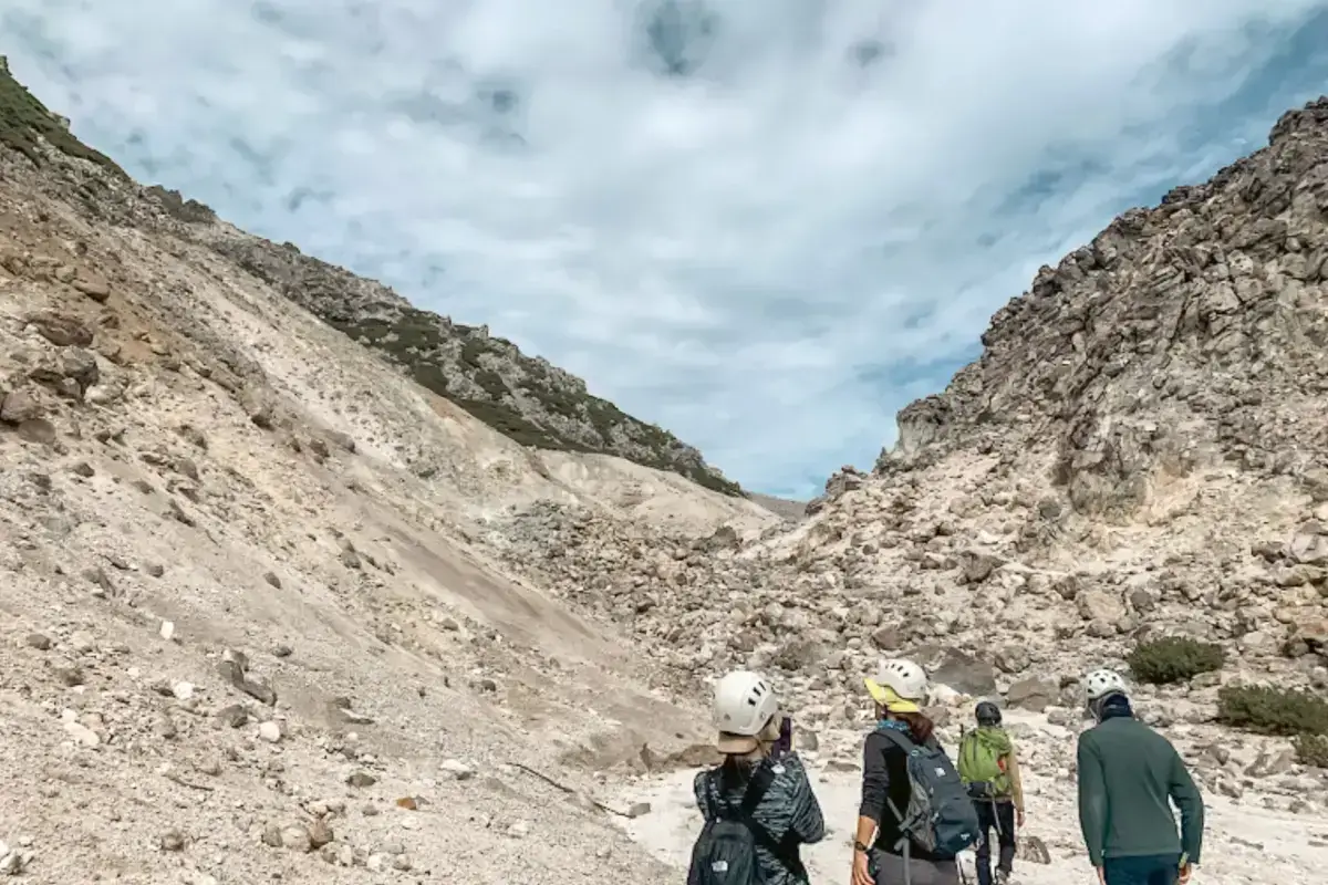Hikers wearing helmets traverse a volcanic rocky gorge with steep barren slopes rising on both sides in Teshikaga, Hokkaido.