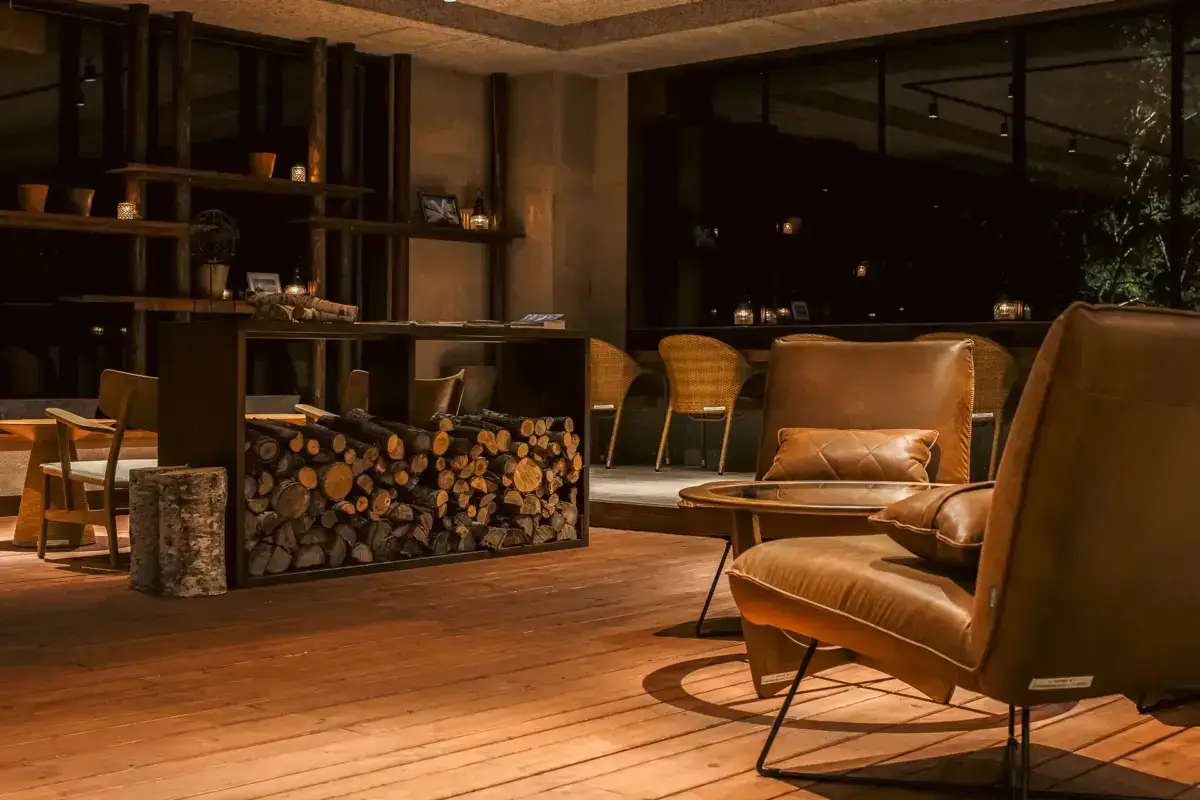 Cozy modern lounge in Teshikaga, Hokkaido featuring tan leather armchairs, stacked firewood, wooden shelving, and floor-to-ceiling windows overlooking a night forest.