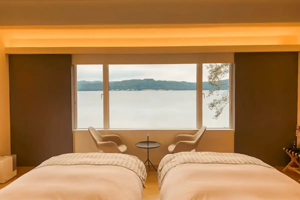 Modern Japanese hotel room with two beds and panoramic windows overlooking Lake Kussharo and forested mountains in Teshikaga, Hokkaido.