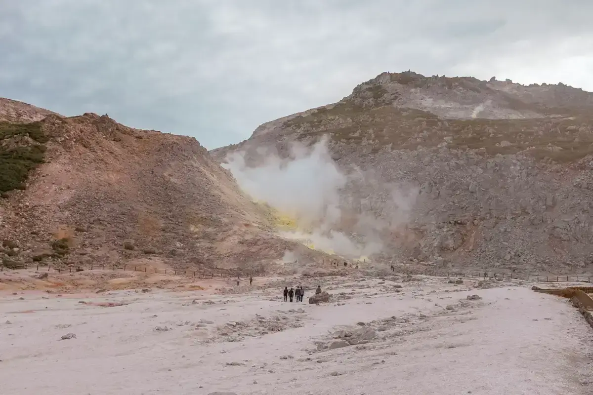 Visitors walking through a volcanic geothermal landscape with sulfurous steam vents rising from rocky hills in Teshikaga, Hokkaido.