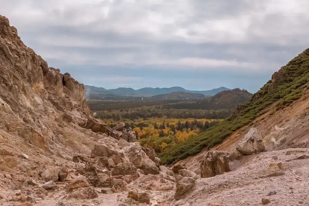 Rugged volcanic rock formations frame a panoramic view of autumn-colored forests and distant mountain ranges in Teshikaga, Hokkaido, under a dramatic cloudy sky.