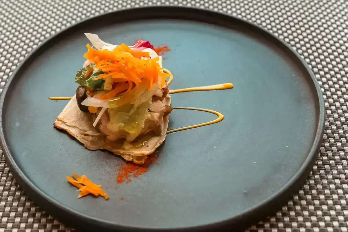 Artfully plated Japanese appetizer on blue ceramic dish featuring crispy crackers topped with pickled vegetables and colorful julienned garnish including carrot and daikon radish, accented with yellow sauce swirl.