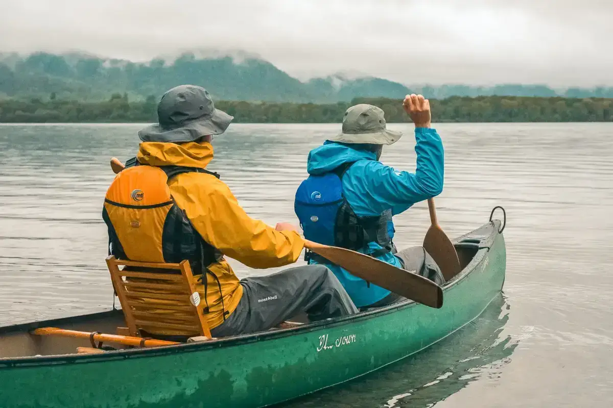 Two people canoeing on a misty lake in Teshikaga, Hokkaido, wearing colorful rain gear and life jackets with forested mountains in the background.
