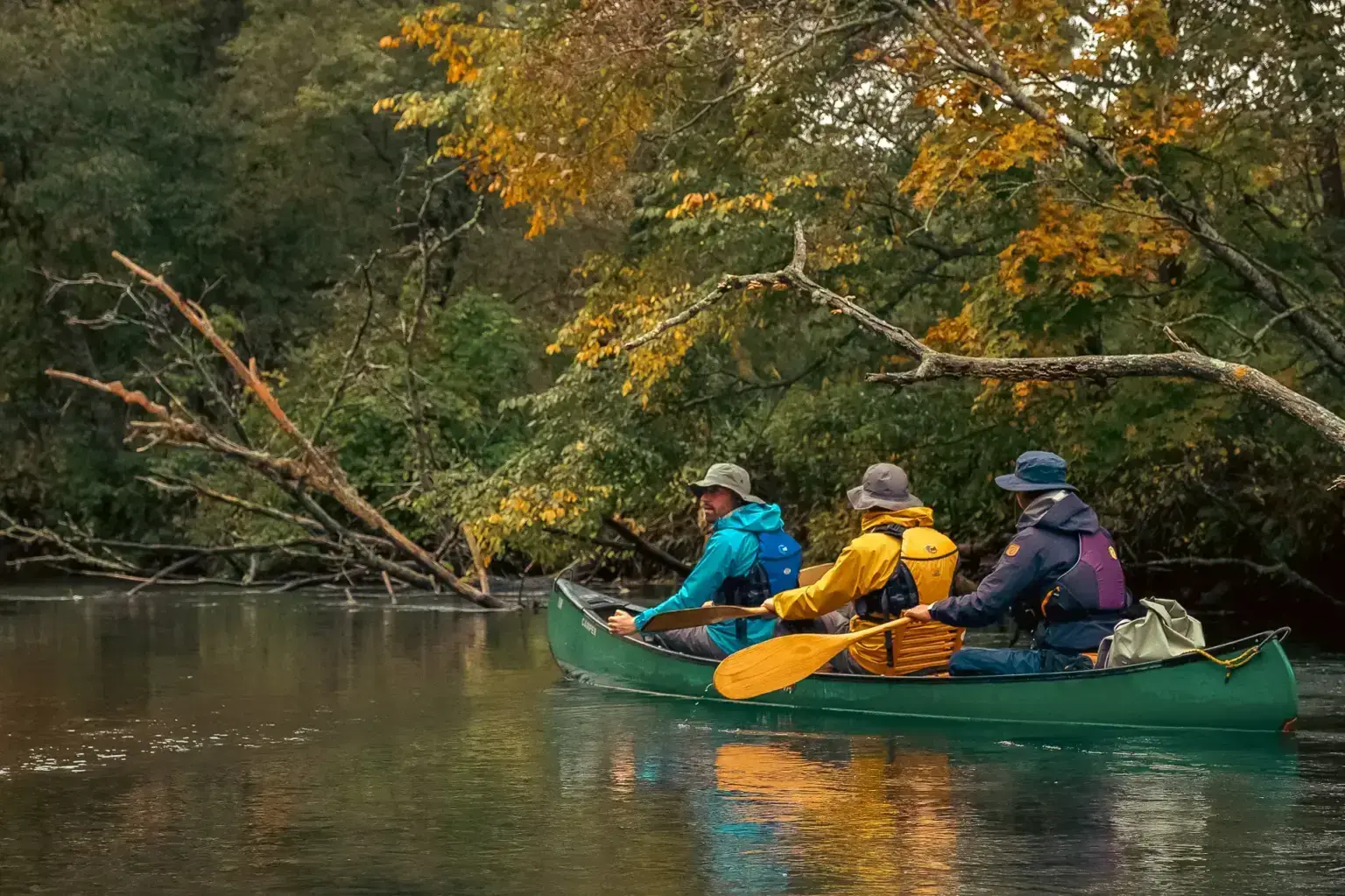 Three people paddling a green canoe on a calm river surrounded by autumn foliage in Teshikaga, Hokkaido.