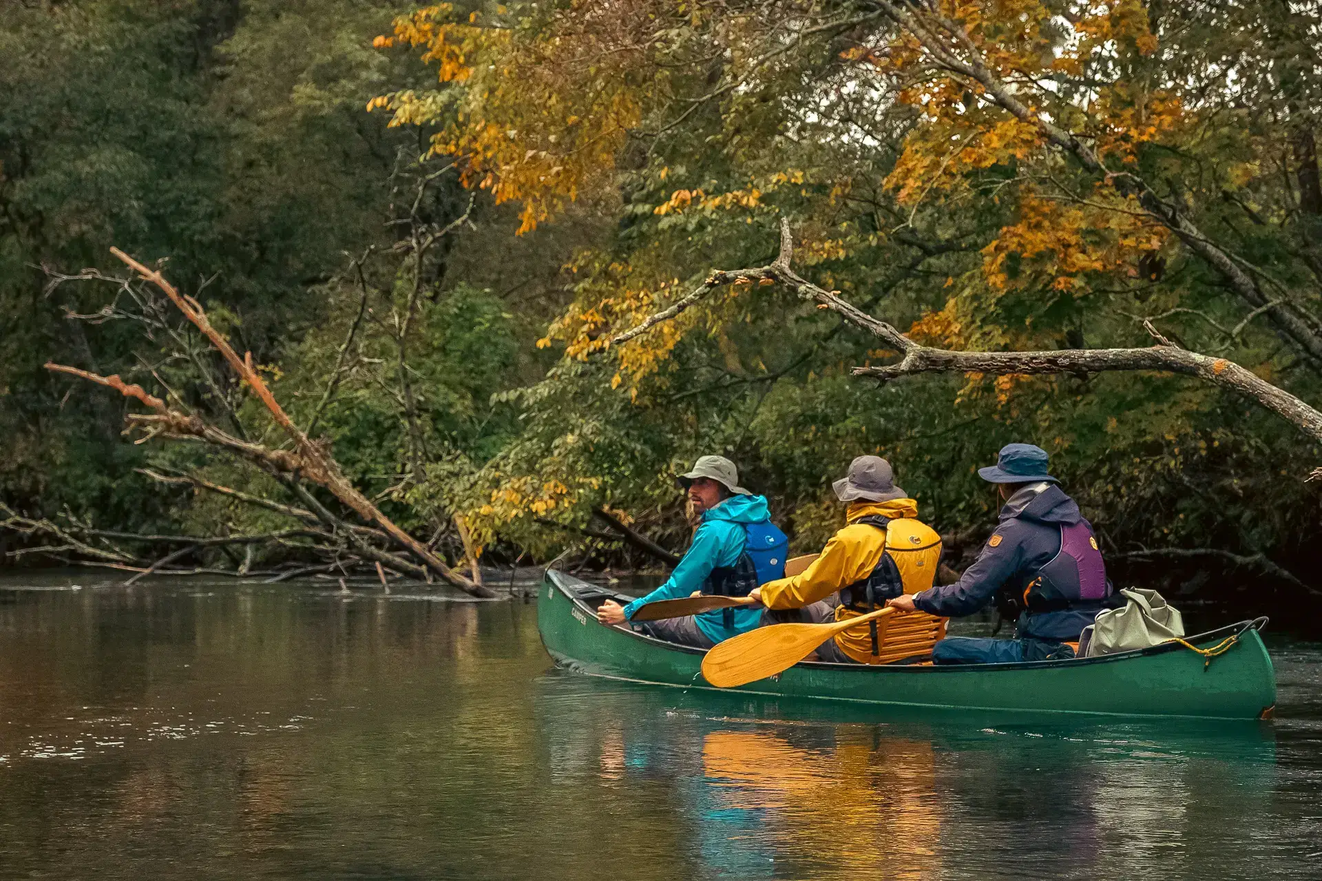 Three people paddling a green canoe on a calm river surrounded by autumn foliage in Teshikaga, Hokkaido.