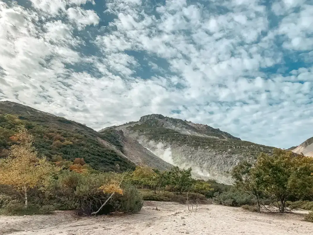 Rocky volcanic mountain with steam vents rising from its slopes, surrounded by low vegetation and autumn foliage under a dramatic cloudy sky in Teshikaga, Hokkaido.