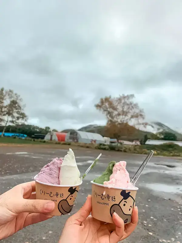 Two hands holding soft-serve ice cream cups with purple, white, and matcha green flavors against a backdrop of farm greenhouses and mountains in Teshikaga, Hokkaido on an overcast day.