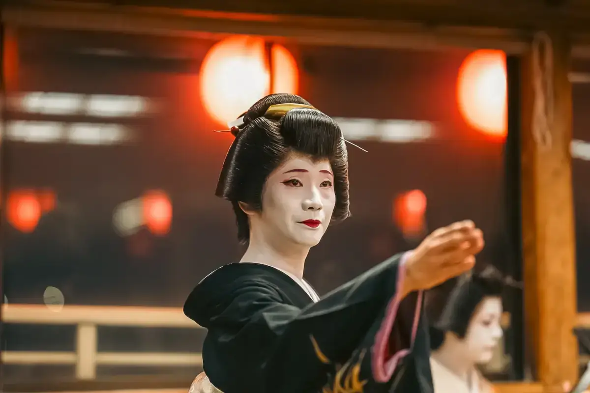 Geisha in traditional black kimono with white makeup and ornate hairstyle performing gracefully in front of illuminated red lanterns at a Japanese temple or traditional venue.