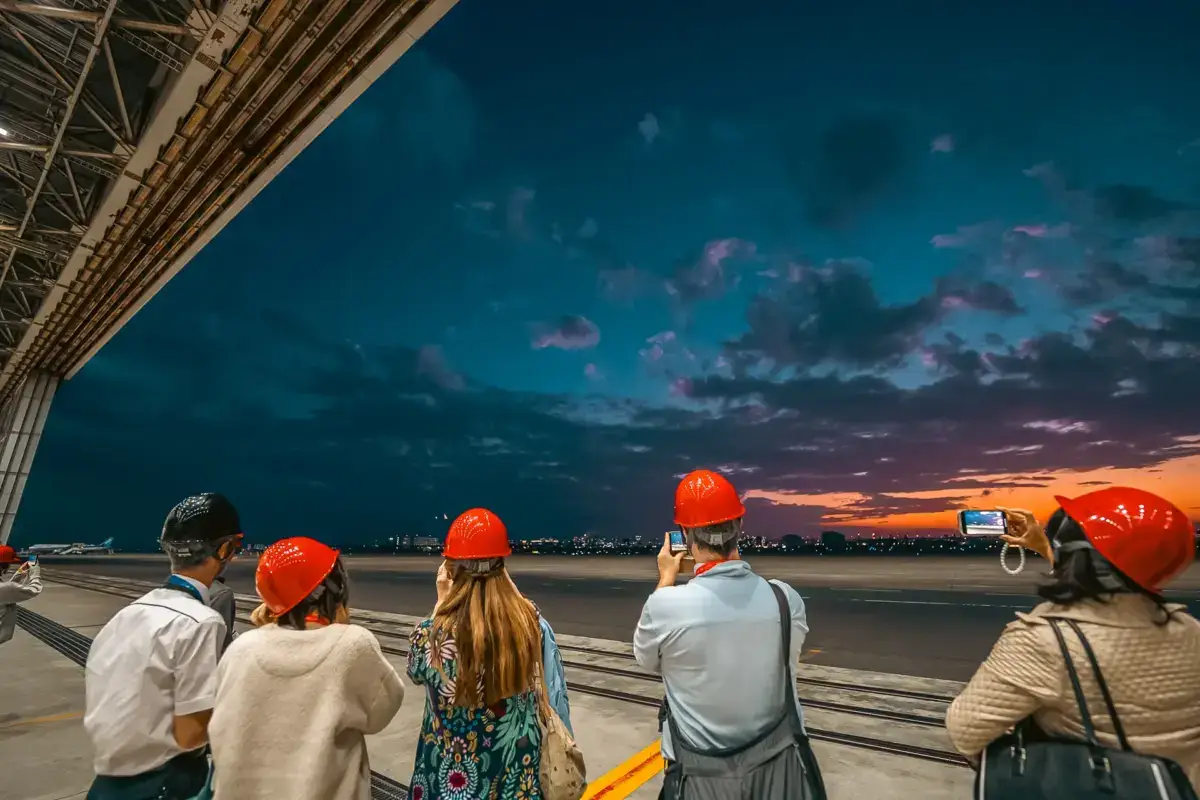 Tourists in bright orange hard hats observe Haneda Airport runway at dusk from an observation deck, with dramatic blue and orange twilight sky overhead.