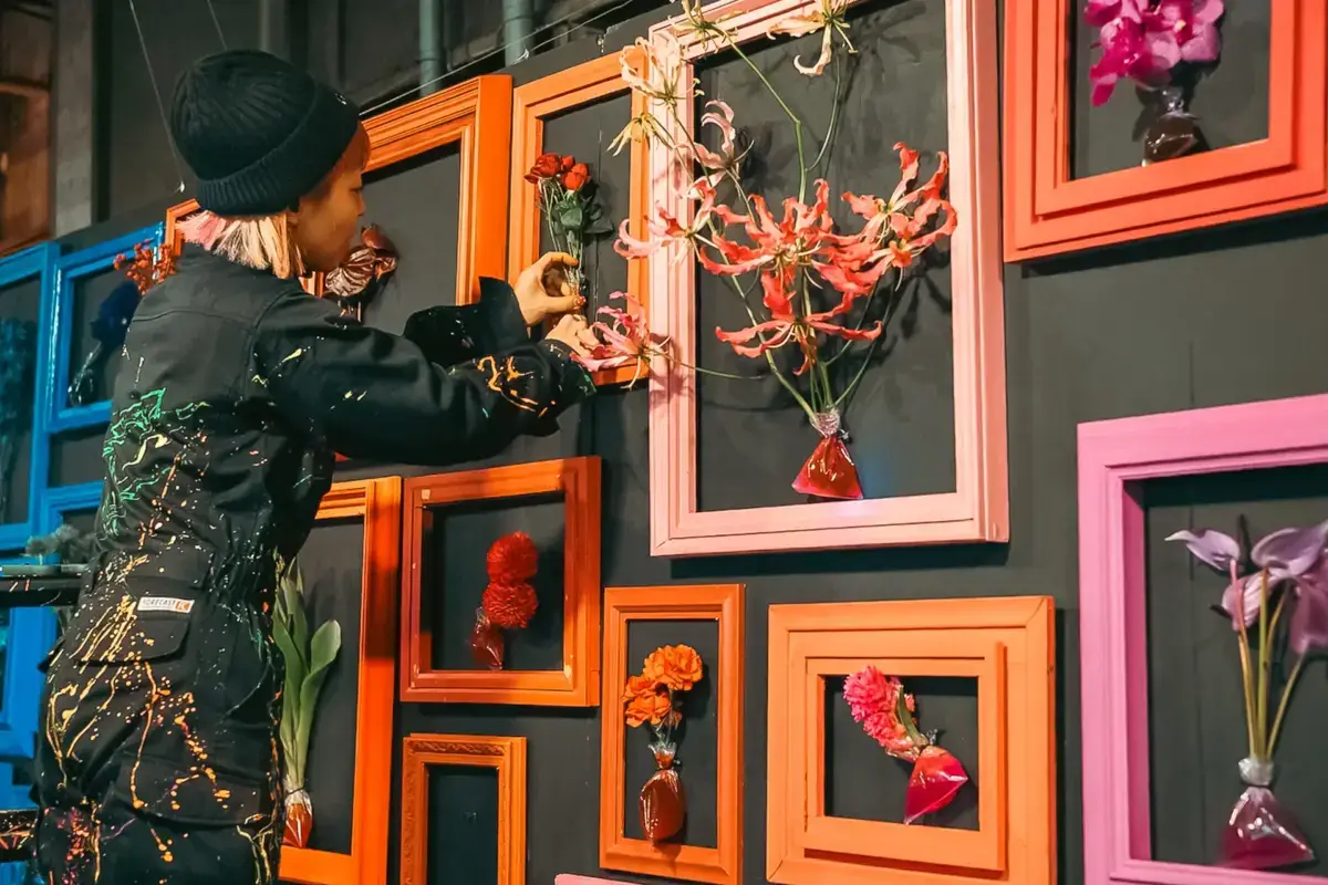 Woman in black winter outfit and knit hat admiring a colorful gallery wall featuring vibrant orange and pink frames with displayed flowers against a dark background in Ota, Tokyo.