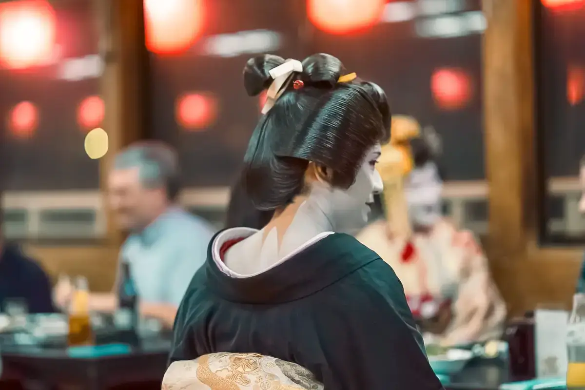 Geisha in traditional attire with elaborate black hairstyle and white makeup, wearing a dark green kimono with cream obi, viewed from behind in a festive setting with red lanterns.
