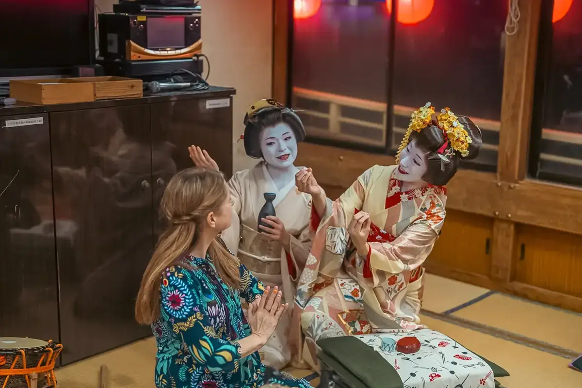 Two geisha in traditional kimono and ornate hair accessories engage with a Western visitor in a colorful yukata during a cultural experience in a traditional Japanese tatami room.