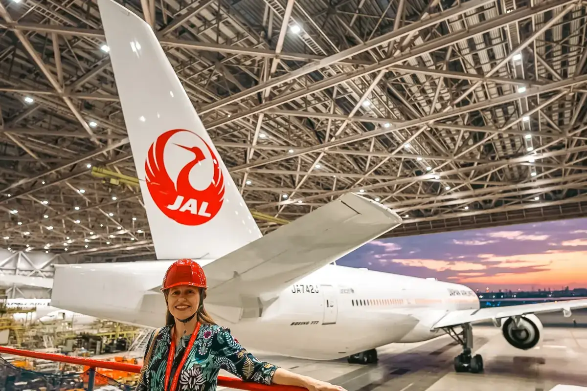 Woman wearing an orange hard hat standing in front of a Japan Airlines Boeing 777 aircraft inside a maintenance hangar with exposed steel framework ceiling.