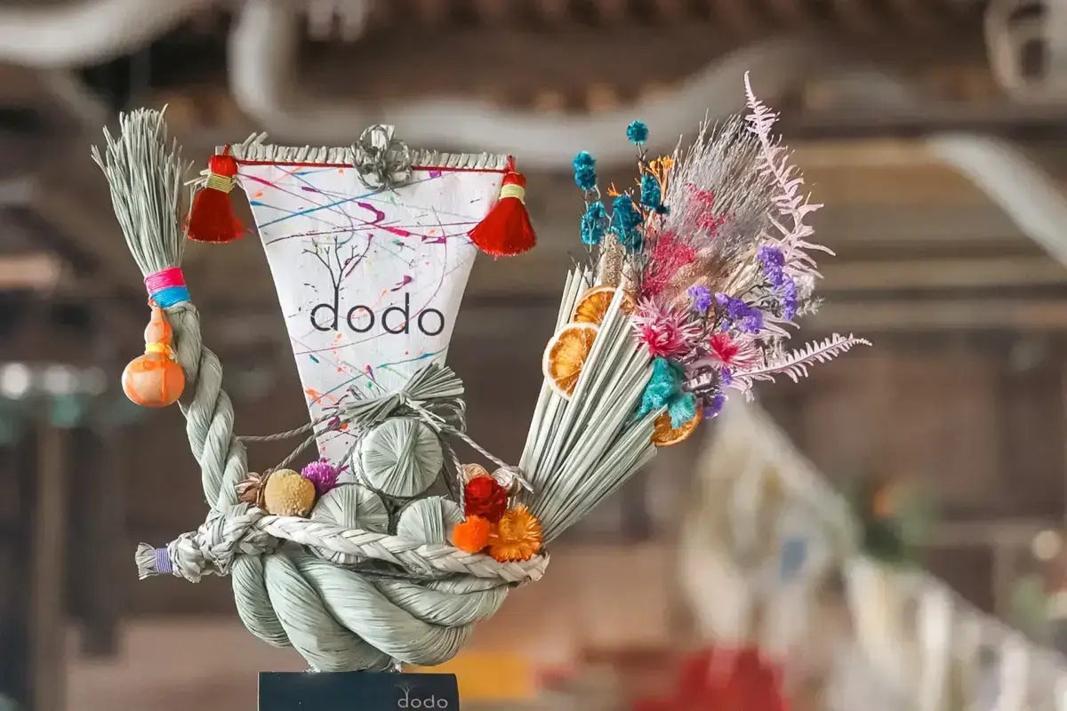 Handcrafted New Year decoration made from twisted straw rope adorned with dried flowers, orange slices, red tassels, and colorful pom-poms with a paper label reading "dodo" at its center.