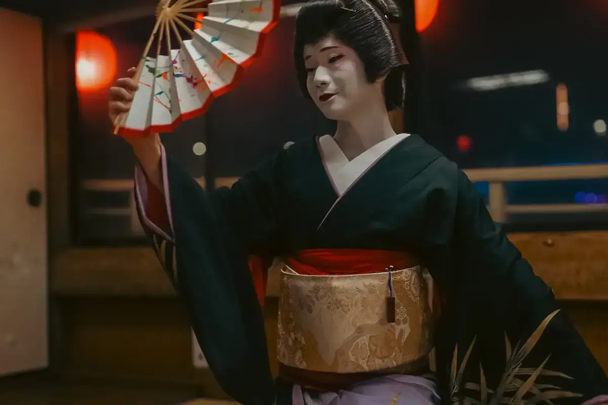 Geisha in traditional dark green kimono and white makeup holding a decorative paper umbrella with red and white pattern, wearing an ornate golden obi in Ota, Japan.