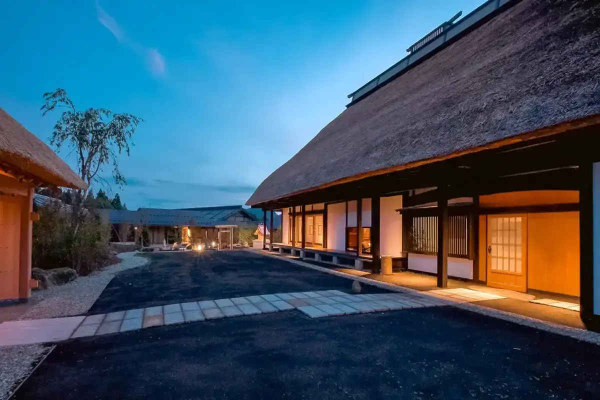Traditional Japanese thatched-roof buildings illuminated warmly at dusk, featuring sliding shoji doors and a gravel courtyard with a willow tree against a blue twilight sky.