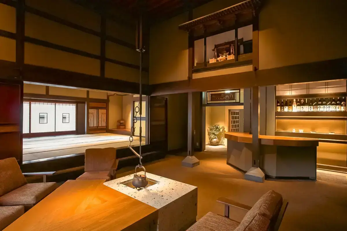 Traditional Japanese machiya interior featuring an open lounge area with exposed wooden beams, a central irori hearth with hanging kettle, ambient lighting, and a bar stocked with sake bottles in the background.