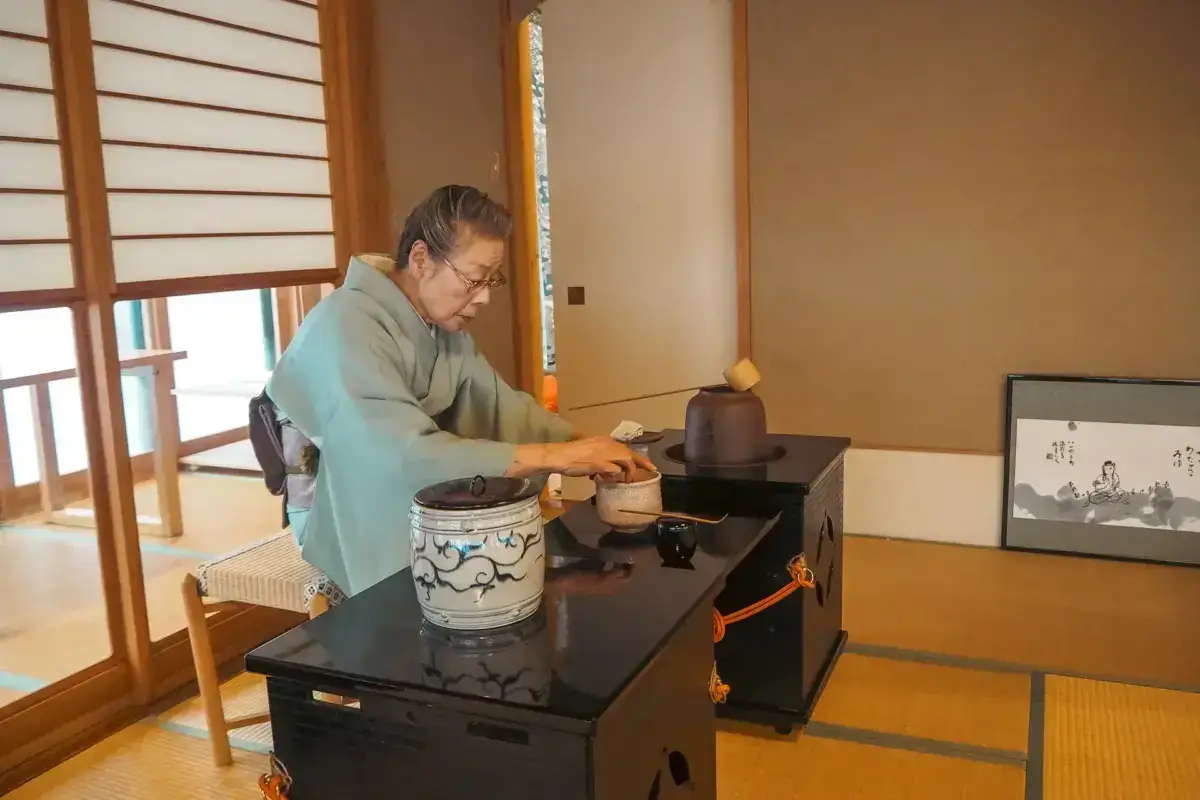 Japanese woman in traditional light blue kimono performing a tea ceremony in a tatami room with shoji screens, carefully preparing matcha tea using traditional ceramic vessels and utensils.