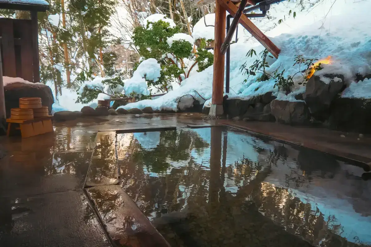Traditional Japanese outdoor onsen hot spring bath surrounded by snow-covered rocks and trees, with wooden buckets and atmospheric steam rising in a winter setting.