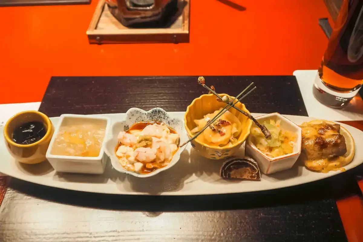 Elegantly arranged kaiseki appetizer course featuring assorted small dishes including shrimp, grilled items on skewers, and seasonal preparations served on a white rectangular plate against a vibrant orange table setting.