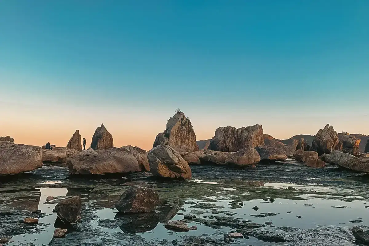 Dramatic coastal rock formations rising from shallow tidal waters at sunset, with jagged spires silhouetted against a clear blue and orange sky along the Japanese coastline.
